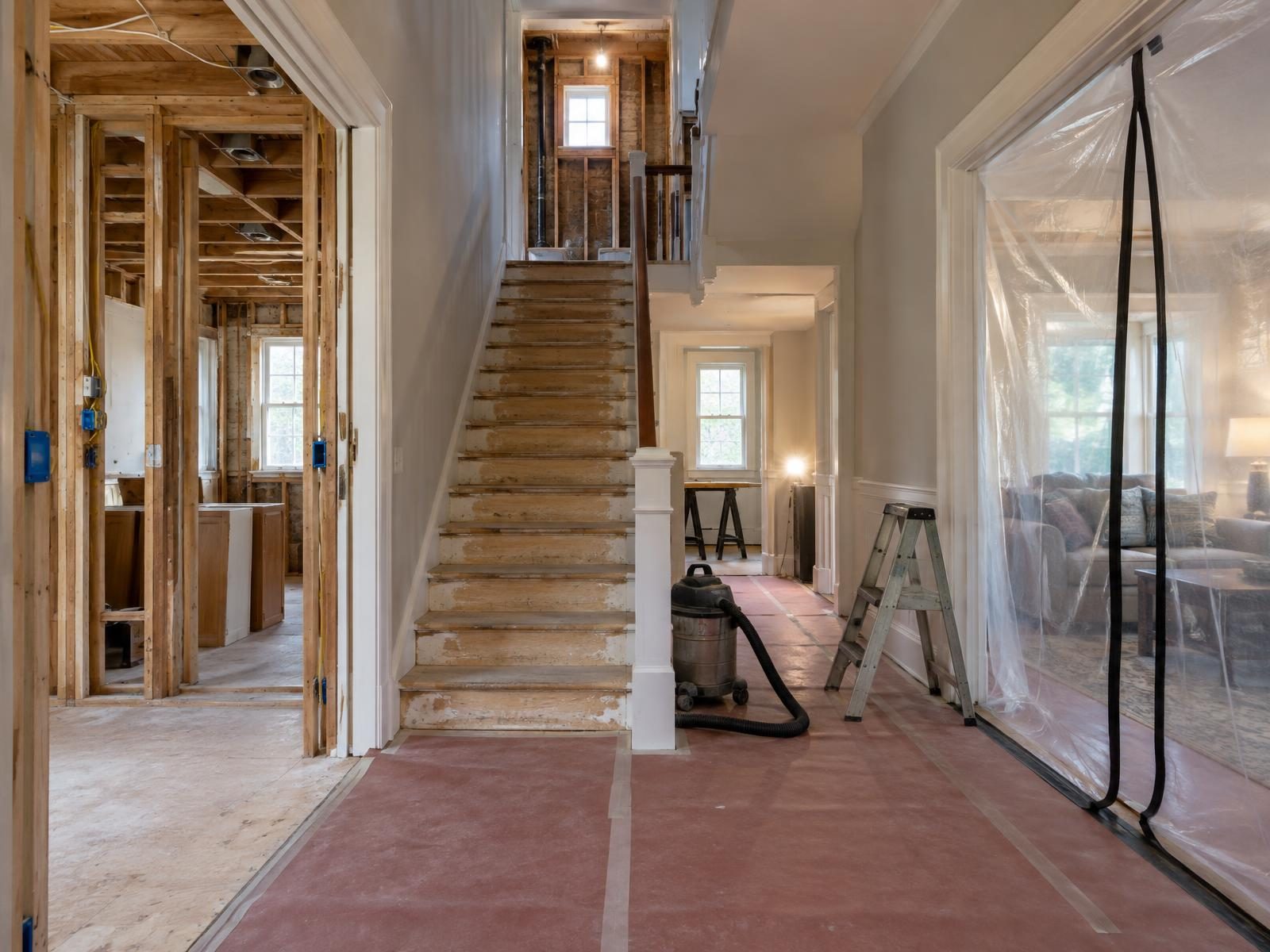 Illustration of a two-story house in the middle of a whole-house renovation, with the kitchen stripped to the studs, a gutted upstairs bathroom, new wiring in open walls and plastic sheeting sealing off the rooms still in use.