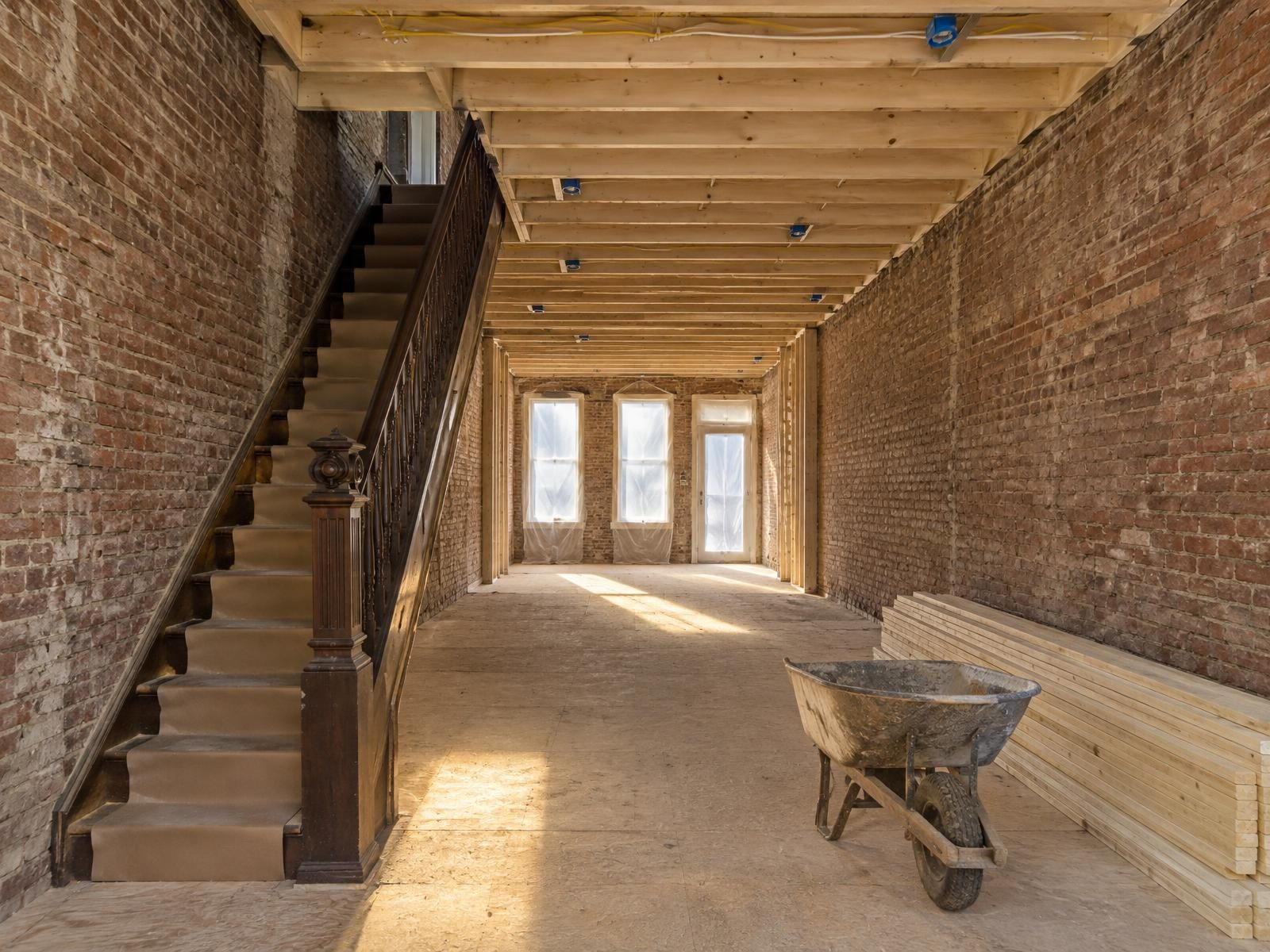 Illustration of a Baltimore rowhouse gutted from front to back during a whole-house renovation, with bare brick party walls on both sides, new joists and wiring overhead, the original staircase left in place and plastic sheeting over the front door.