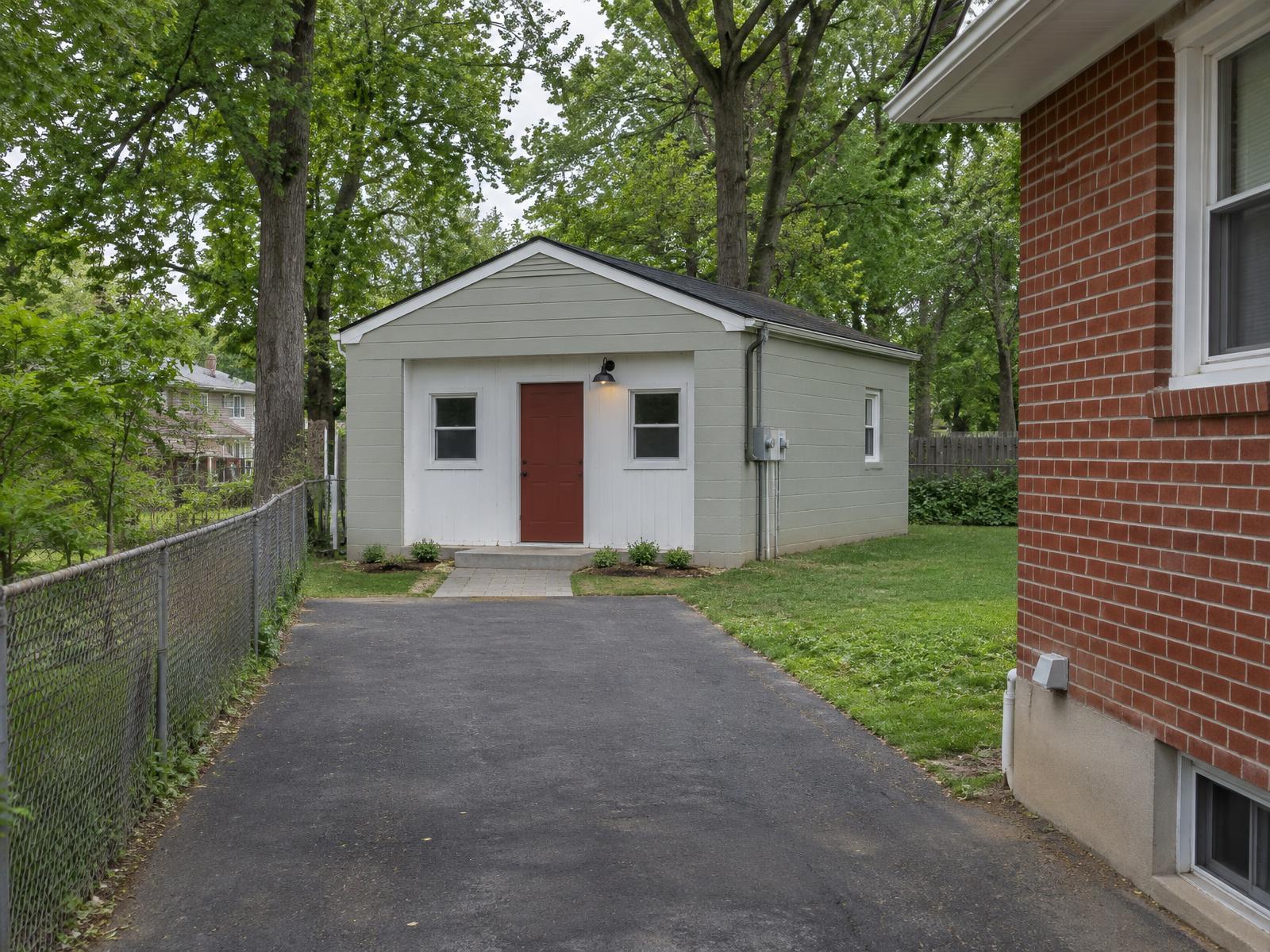 Illustration of a 1950s brick rambler in Camp Springs whose detached garage has been converted into a one-bedroom backyard cottage, with a new entry door and windows where the garage door was, a paved path from the driveway and a porch light.