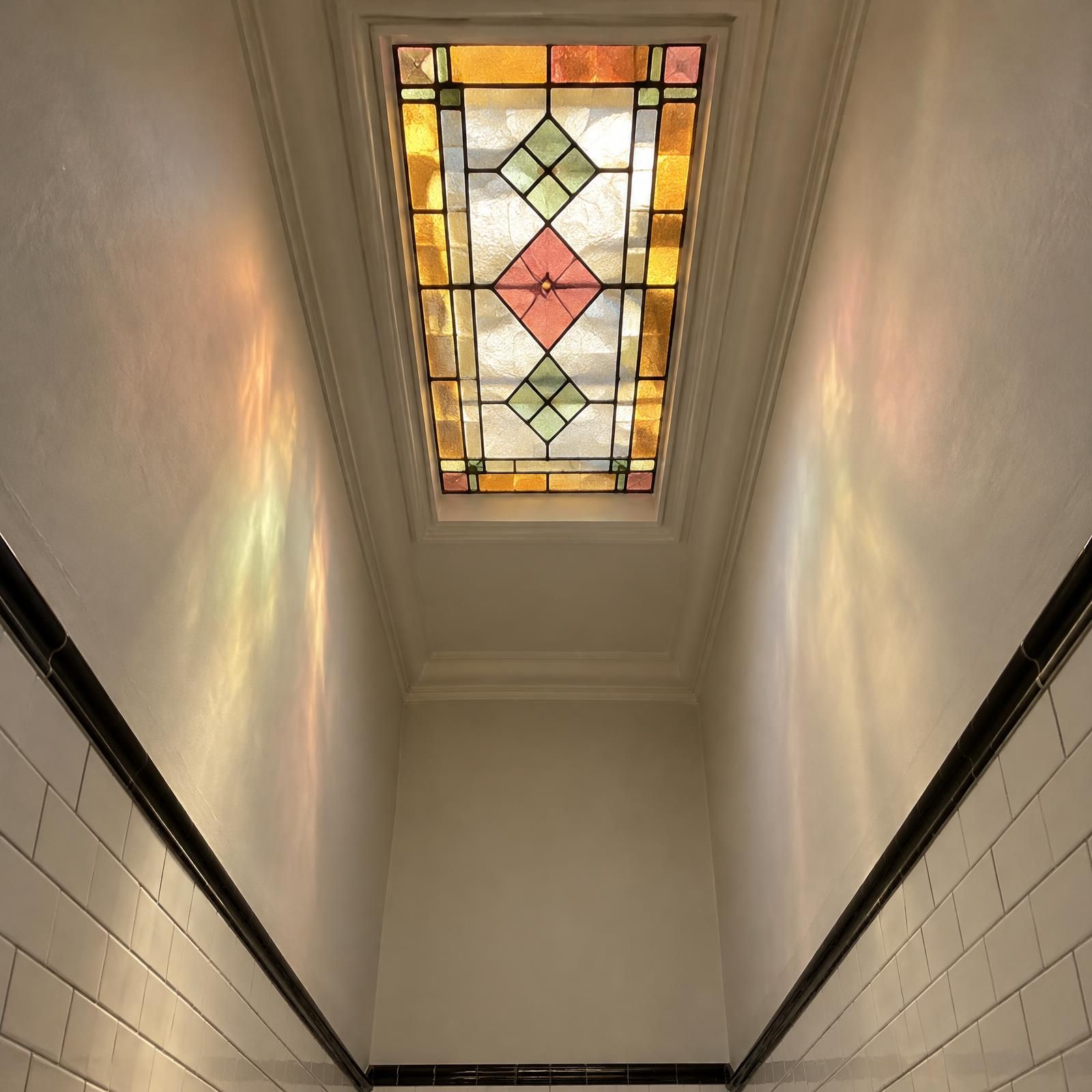 Illustration of a stained-glass skylight above a long, narrow rowhouse bathroom with white tile on the lower walls.