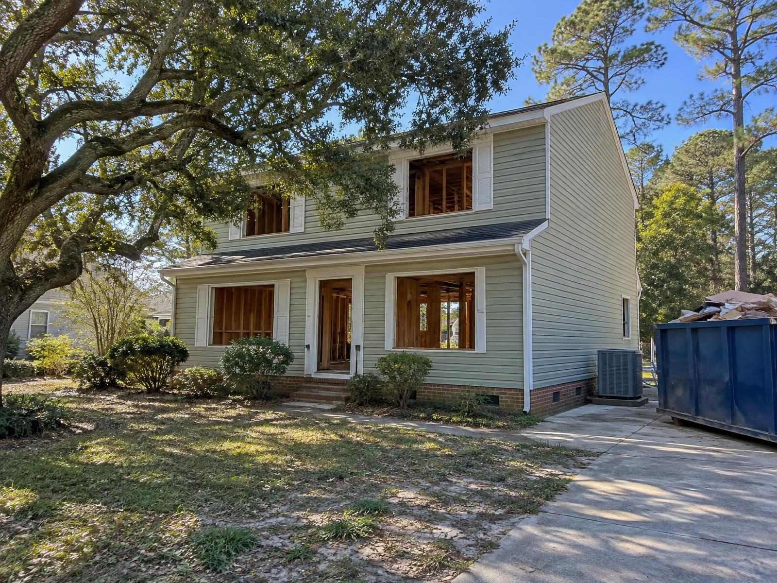 Illustration of a 1980s two-story Virginia Beach house with vinyl siding in the middle of a whole-house renovation, the downstairs opened to the studs, a new heat pump on a raised pad beside the foundation and a dumpster in the driveway.